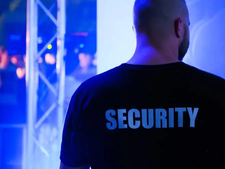 A door supervisor in a black Security shirt at a venue under blue stage lighting